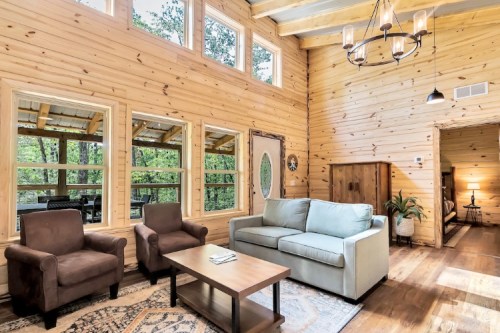 Cozy wooden living room with large windows, a sofa, two armchairs, and a coffee table, surrounded by nature.