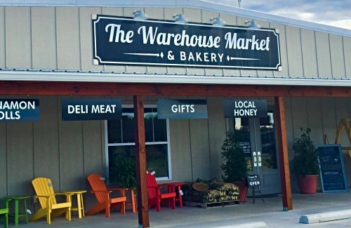 Exterior of The Warehouse Market & Bakery, featuring colorful chairs and signs for deli meat, gifts, and local honey.