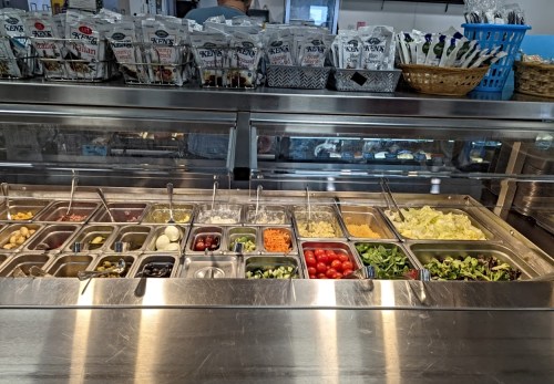 A salad bar with various toppings, including vegetables, olives, and dressings, in a stainless steel display.