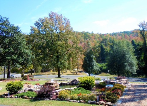 A scenic view of a landscaped yard with colorful flowers, trees, and a backdrop of autumn foliage on rolling hills.