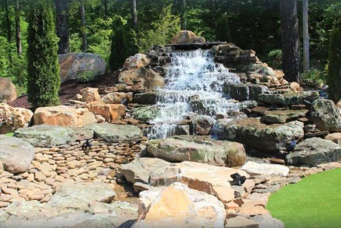 A serene waterfall cascading over rocks, surrounded by lush greenery and natural stone landscaping.