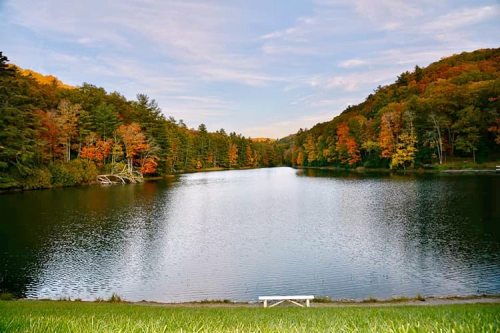 A serene lake surrounded by colorful autumn trees under a clear sky, with a bench in the foreground.