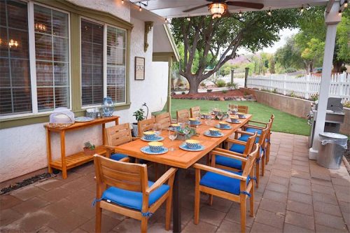 A spacious outdoor dining area with a long wooden table set for a meal, surrounded by chairs and greenery.
