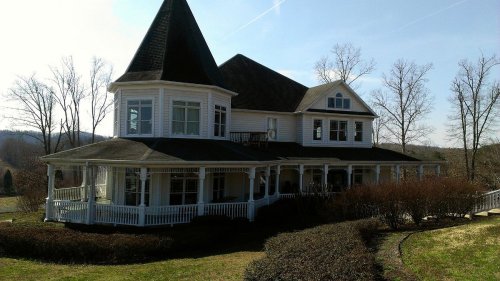 A large, white Victorian-style house with a pointed roof and wraparound porch, surrounded by trees and shrubs.