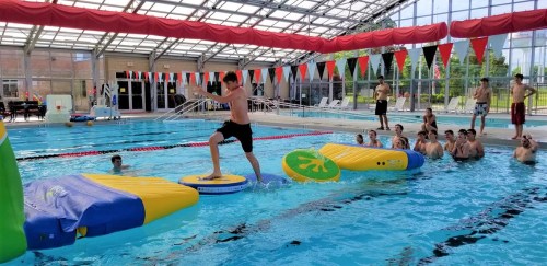 A boy balances on a floating obstacle in a pool while others watch from the side, enjoying a fun water activity.