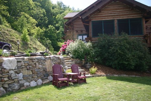 A cozy outdoor area with two red chairs beside a stone wall, lush greenery, and a log cabin in the background.