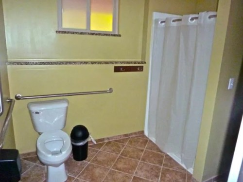 A bathroom featuring a toilet, shower with curtain, trash can, and tiled floor with yellow walls.