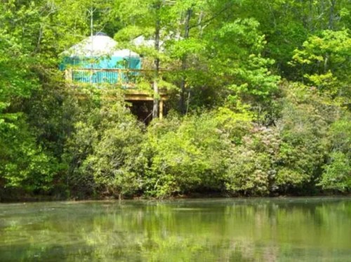 A serene view of a lush green landscape with a wooden deck and a yurt by a calm, reflective pond.