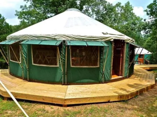 A green yurt with a wooden deck, surrounded by trees in a natural setting.