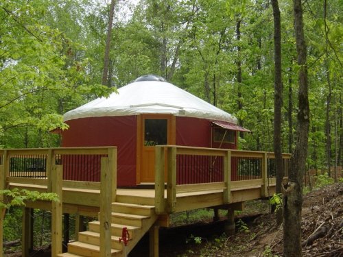 A red yurt on a wooden deck surrounded by lush green trees in a forested area.