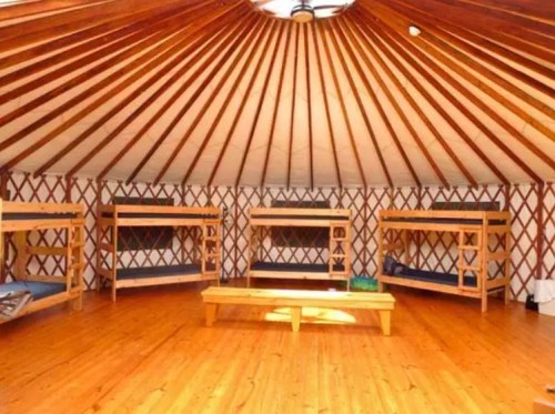 Interior of a yurt featuring wooden bunk beds and a circular ceiling with wooden beams, set on a wooden floor.