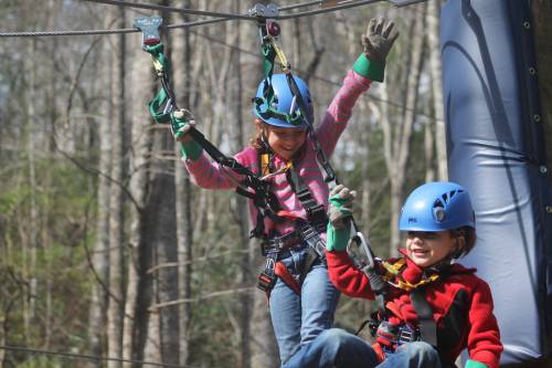 Two children in helmets and harnesses enjoy a zip line adventure in a forested area, smiling and having fun.