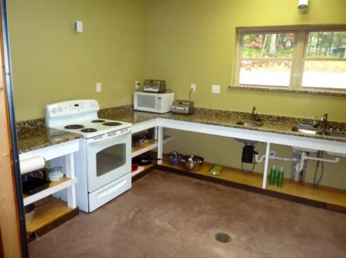 A modern kitchen with a white stove, microwave, sink, and granite countertops, featuring wooden shelves and a concrete floor.