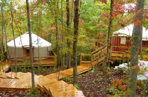 A wooden pathway leads through a forest to two yurts surrounded by colorful autumn foliage.