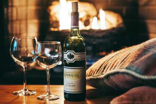 A bottle of Merlot wine with two glasses, set beside a cozy fireplace and a patterned pillow.