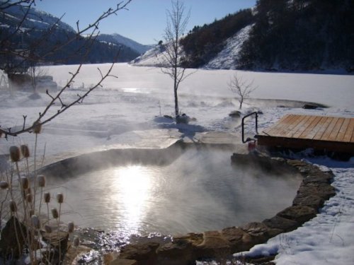 A serene hot spring surrounded by snow, with mist rising and mountains in the background under a clear blue sky.