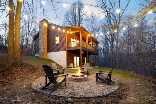 A cozy cabin with string lights, surrounded by trees, featuring a fire pit and seating area in the foreground.