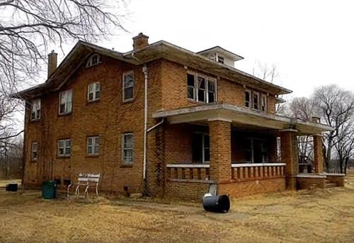 Abandoned two-story brick house with broken windows and a porch, surrounded by overgrown grass and trees.