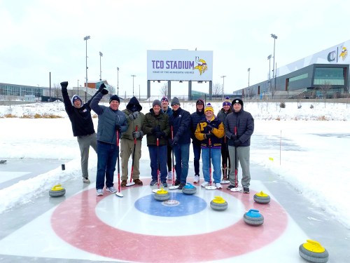 A group of people poses on an outdoor curling rink, smiling and celebrating with curling stones and a stadium in the background.