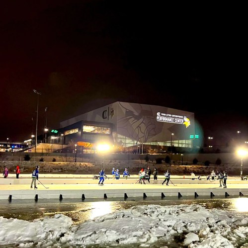 A nighttime scene of an outdoor ice rink with players skating, illuminated by bright lights and a performance center in the background.
