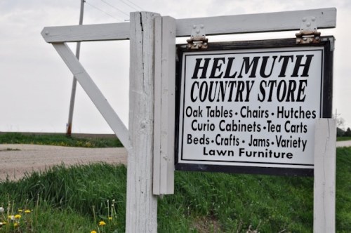 Sign for Helmuth Country Store featuring items like oak tables, chairs, and lawn furniture, set against a rural backdrop.