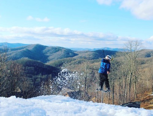 A skier jumps off a snowy ramp, with mountains and a blue sky in the background. Snow sprays into the air.