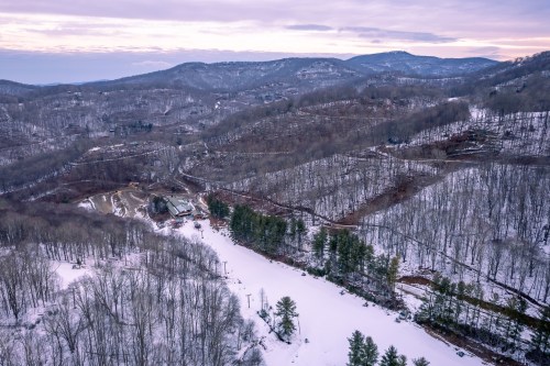 Aerial view of a snow-covered mountainous landscape with bare trees and a winding road.