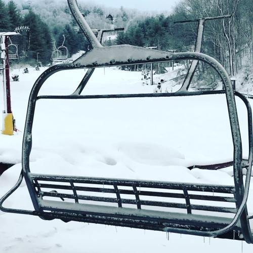 A snow-covered ski lift chair against a winter landscape with trees and a snowy slope in the background.