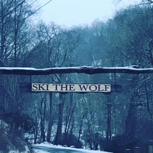 Sign reading "SKI THE WOLF" hanging over a snowy path surrounded by trees.