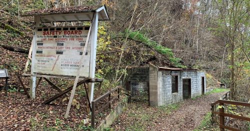 A weathered safety board stands near two abandoned buildings in a wooded area, surrounded by autumn foliage.