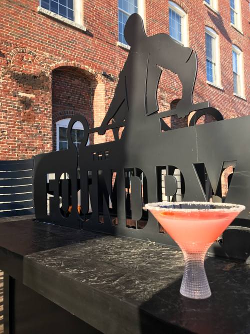 A pink cocktail with a salted rim sits in front of a black sign reading "The Foundry," against a brick wall backdrop.