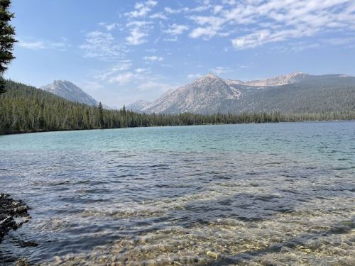 A serene lake surrounded by mountains and lush trees under a clear blue sky. Water is calm and crystal clear.