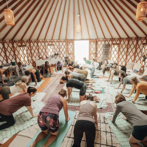A group of people practicing yoga in a spacious, warmly lit yurt with wooden beams and mats on the floor.