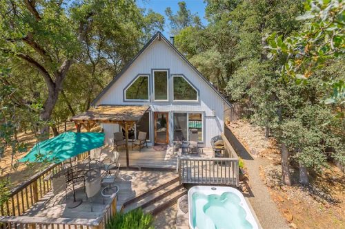 A cozy A-frame cabin surrounded by trees, featuring a deck, outdoor seating, and a hot tub.