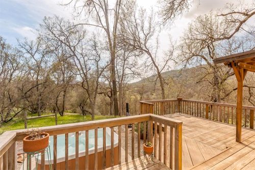 A wooden deck overlooks a serene landscape with trees and a hot tub, under a bright sky.