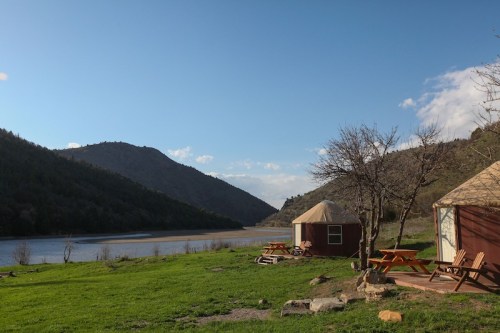 A serene landscape featuring a river, mountains, and two yurts surrounded by green grass and picnic tables.