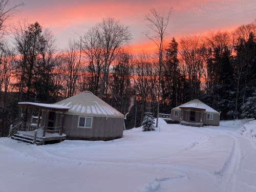 Two yurts surrounded by snow, with a vibrant sunset and trees in the background.