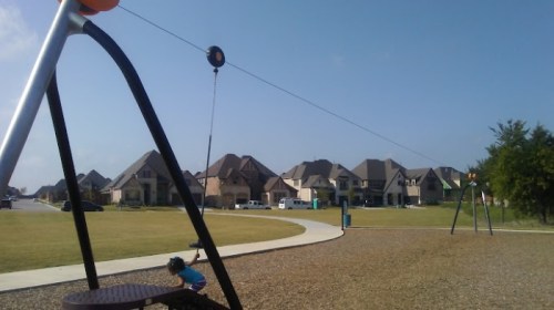 A playground with a swing set and a zip line, surrounded by houses under a clear blue sky. A child plays nearby.