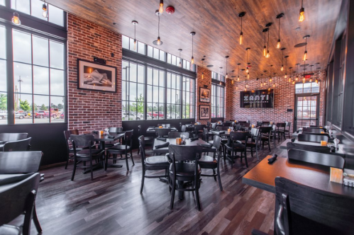 Interior of a modern restaurant featuring wooden floors, brick walls, and large windows with empty tables and chairs.
