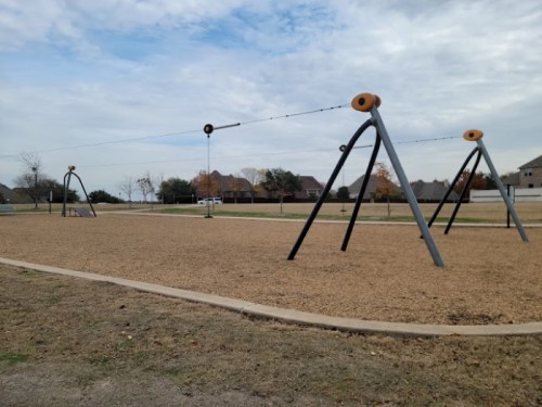A playground with swings and a zip line, surrounded by grass and trees under a cloudy sky.