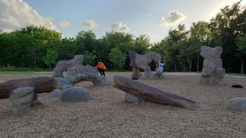 Two children play on a natural-themed playground with rock-like structures and logs, surrounded by trees and a cloudy sky.