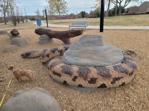 A large, realistic snake sculpture lies on the ground in a park, with a small dog nearby and trees in the background.