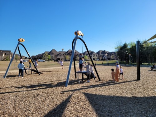 Children playing on swings and a playground in a sunny park with houses in the background.