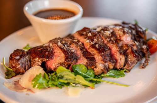 A perfectly cooked steak, sliced and served on a plate with greens and a small bowl of sauce.