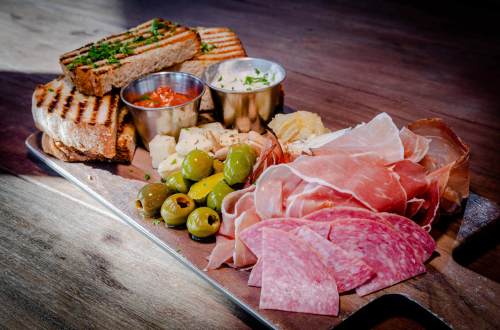 A wooden platter with grilled bread, olives, cured meats, cheese, and dipping sauces.