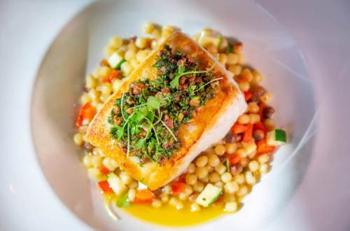 A beautifully plated piece of fish topped with herbs, served on a bed of couscous and colorful diced vegetables.