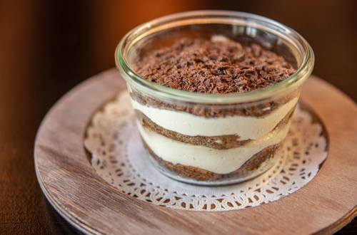 A glass dessert dish layered with cream and chocolate shavings, resting on a wooden plate with a lace doily.