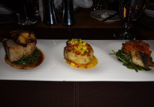 Three gourmet dishes on a white plate: grilled fish with sauce, a mango-topped crab cake, and a fish fillet with tomato.