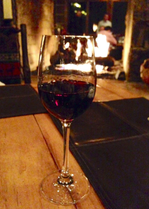 A glass of red wine sits on a wooden table, with a warm fireplace glowing in the background.