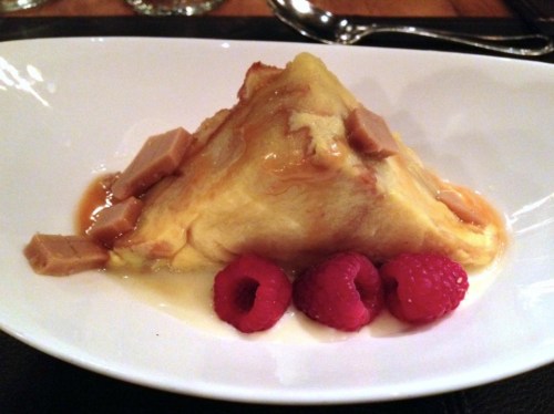 A triangular dessert topped with caramel sauce and pieces of fudge, accompanied by fresh raspberries on a white plate.